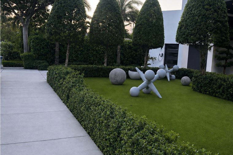 A manicured front garden with cone-shaped topiary trees, a low clipped hedge, artificial turf and white sculptural spheres along a concrete walkway.
