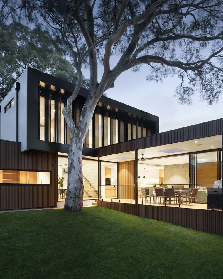 Contemporary home lit from within at dusk, opening onto a wide manicured lawn under a mature shade tree.